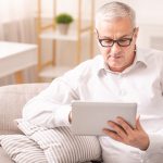 Reading News. Senior man in spectacles using digital tablet sitting on the couch at home, copyspace