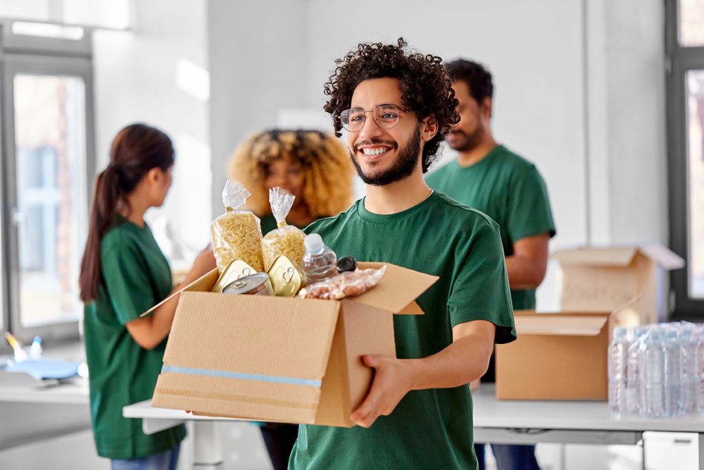 Charity,,Donation,And,Volunteering,Concept,-,Happy,Smiling,Male,Volunteer - WebIXI charity, donation and volunteering concept - happy smiling male volunteer with food in box and international group of people at distribution or refugee assistance center