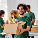 charity, donation and volunteering concept - happy smiling male volunteer with food in box and international group of people at distribution or refugee assistance center